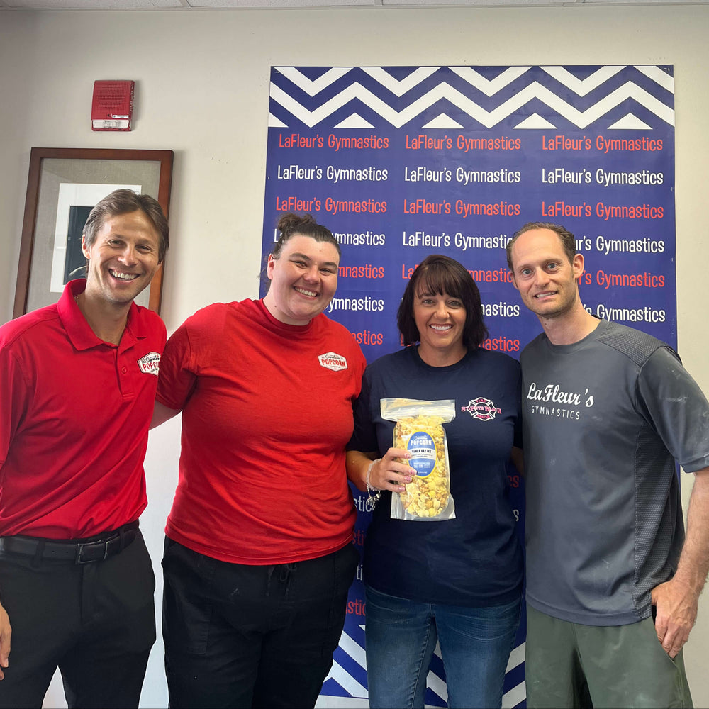 Four people standing together in an indoor office or gym setting, wearing branded LaFlour's Gymnastics shirts and logos, with a blue and white banner behind them and a bag of snacks in the center.
